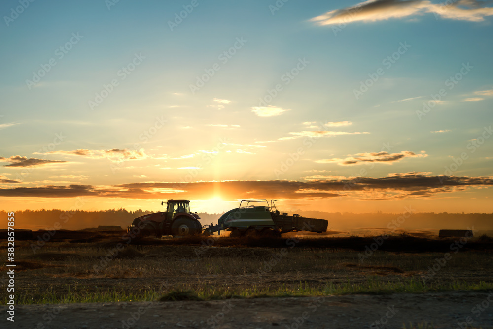 Silhouette of working tractor in autumn field in sunset time, tractor packing hay into rectangular bale, working machine in cloudy golden sky background, agriculture concept