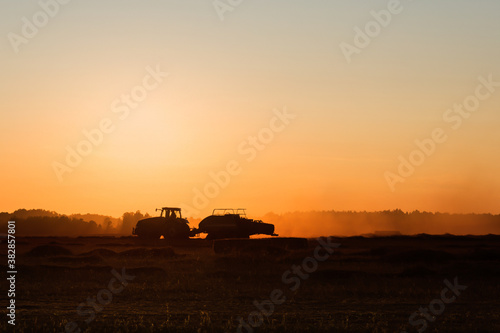 Silhouette of working tractor in autumn field in sunset time  tractor packing hay into rectangular bales  working machine in golden sky background  agriculture concept
