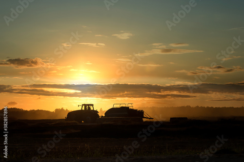 Silhouette of working tractor in autumn field in sunset time, tractor packing hay into rectangular bale, working machine in cloudy golden sky background, agriculture concept
