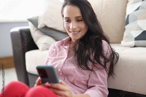 Woman sit on floor near sofa, look at screen and smile. Female hand hold phone at home.