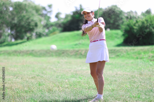 Beautiful woman in white skirt, cap and pink t-shirt play golf on green lawn. Close-up hitting ball with club.