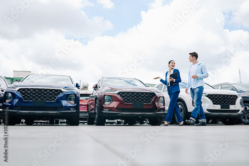 Positive female car dealership saleswoman walking along rows of new cars with customer photo