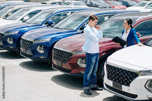 Pretty confident saleswoman showing car to pensive doubting young customer