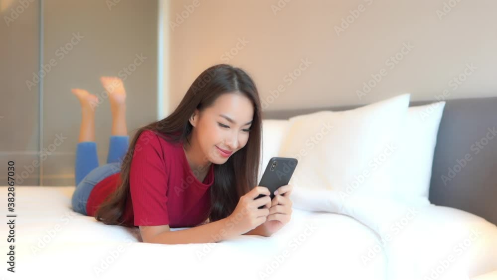 Face Close-up on Asian woman typing a message on a mobile phone using 2 hands while comfortably lying on a bed in a hotel room Face Close-up on Asian woman typing a message on a mobile phone using 2 hands while comfortably lying on a bed in a hotel room