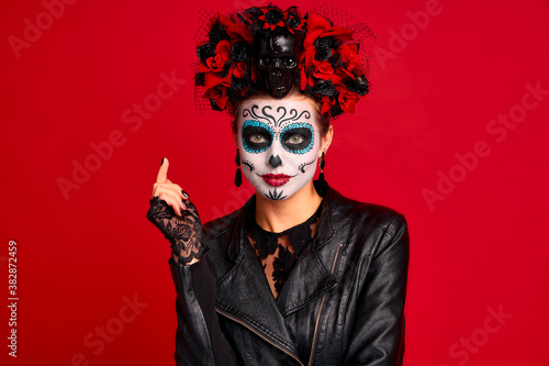 Scary young creepy lady calavera. wears artistic make-up for the feast of all the dead.Wears black leather jacket and lace gloves, dressed as skeleton isolated in red.