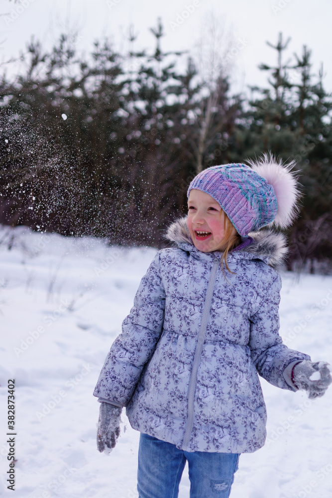 girl in a blue jacket and a fluffy hat plays in the snow in the forest. sincere joy of the child from playing in the fresh air. raising immunity in winter. playing snowballs in the winter forest