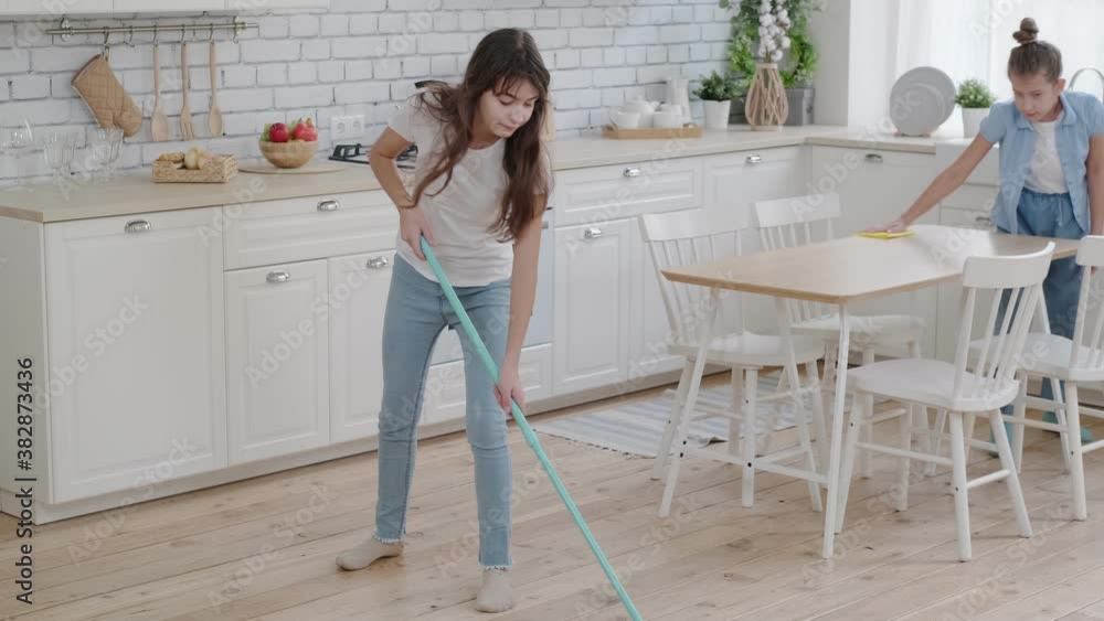 Children are busy with household chores. Little sisters mop floors and dust in the kitchen Children are busy with household chores. Little sisters mop floors and dust in the kitchen