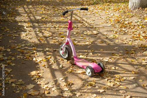 A children's scooter with a large wheel in front of red-pink color stands on the track. There are a lot of yellow dry leaves around. Autumn