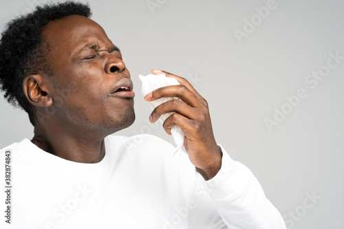 Close up of unhealthy Afro-American man blowing nose and sneeze into tissue or napkin, experiences allergy symptoms, closed eyes, standing over gray background. First symptoms of a cold and flu.  photo