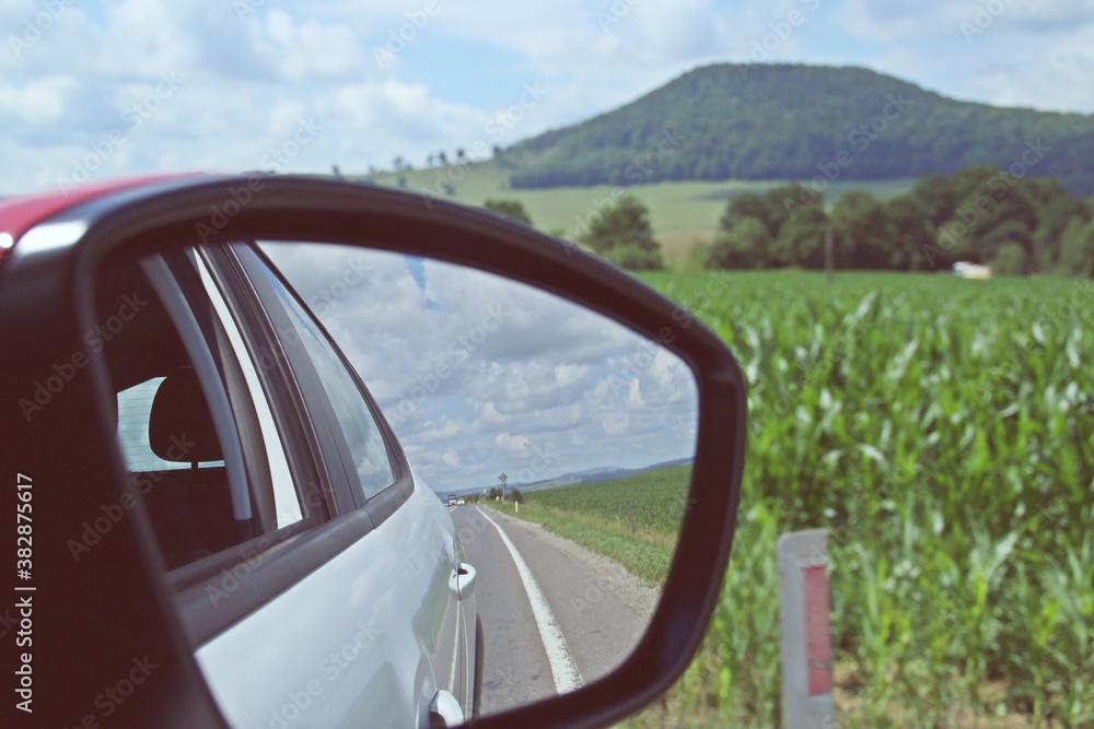 Espejo retrovisor del coche con el reflejo del lateral del mismo. Conduciendo por la carretera E60 entre los campos de cultivo de Transilvania, Rumanía.