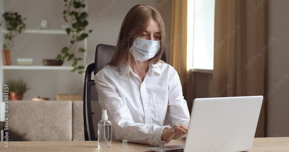 Portrait of business woman sitting at table at home in room or office in company, wears protective medical mask on her face, types on keyboard, works in laptop online, studies remotely looks at camera Portrait of business woman sitting at table at home in room or office in company, wears protective medical mask on her face, types on keyboard, works in laptop online, studies remotely looks at camera