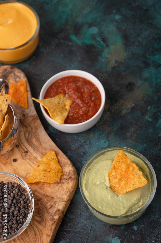 Some corn chips on wooden chopped board with salsa, cheese dip, avocado sous and bowl with spice on dark blue background. Tasty food. Unhealthy meal. Vertical format. Traditional mexican cuisine.