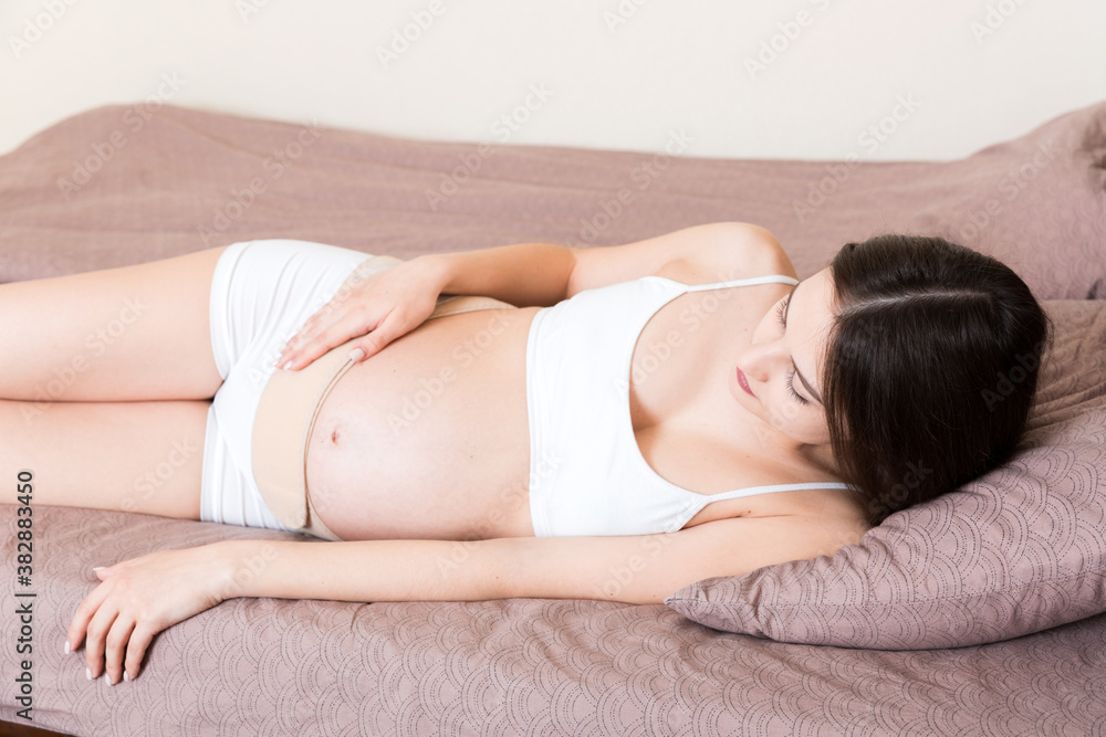 closeup pregnant woman dresses bandage on belly at home on bed. Orthopedic abdominal support belt closeup pregnant woman dresses bandage on belly at home on bed. Orthopedic abdominal support belt