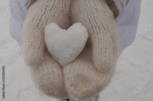Human hands in warm white knitted mittens with a snow heart on a snowy background, romantic Valentine's day concept