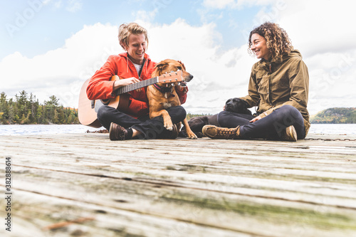 Happy young couple playing with their dog and guitar on a dock - Man and woman in love near a lake in Canada in autumn - love and lifestyle in the nature