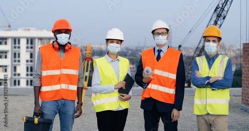 Portrait of multi ethnic group of males and female in helmets and medical masks standing at construction side on top of building and posing to camera. Mixed-races men and woman. Team of builders.