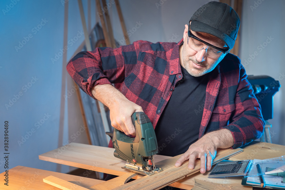 A man in goggles saws a wooden bar. Joiner works with an electric jigsaw. Carpentry tools in the workshop. Woodworking is a hobby. Manufacture of wooden products.