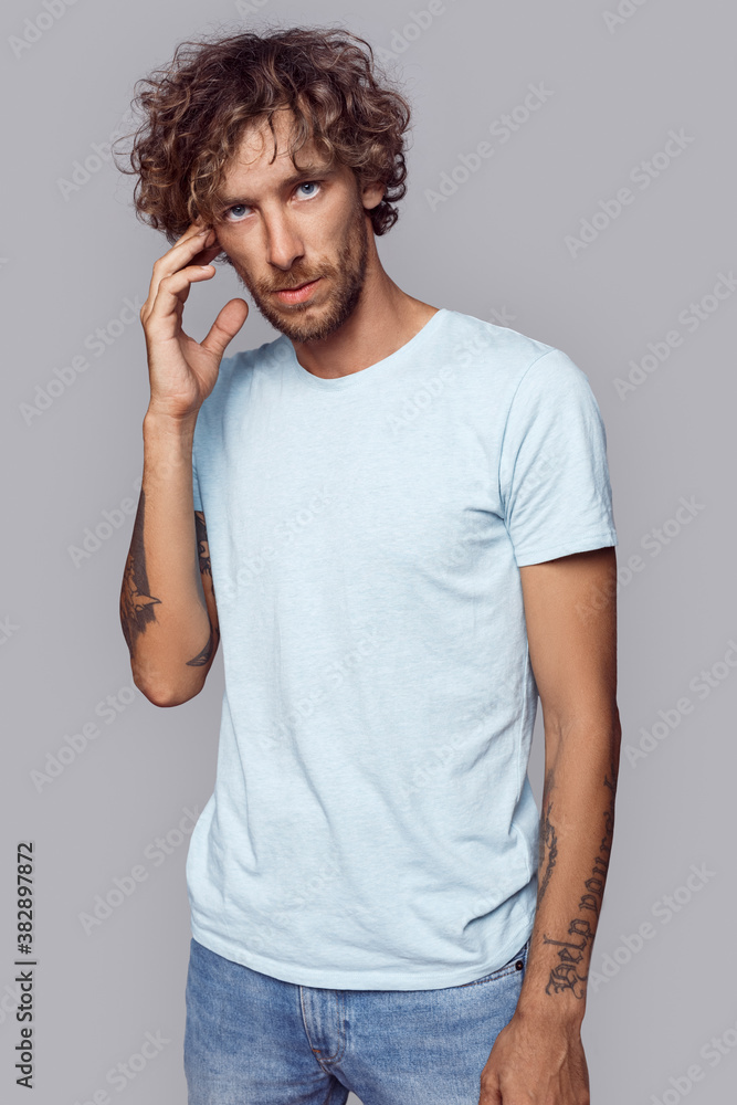 Freestyle. Man with curly hair in t-shirt standing isolated on grey touching head pensive Freestyle. Man with curly hair in t-shirt standing isolated on grey touching head pensive