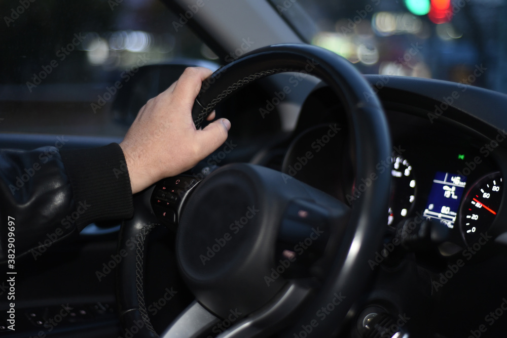 A man's hand holds the steering wheel of a car. The car is coming. The speedometer shows the speed of movement. Behind the glass, you can see the evening street light. Bokeh background.