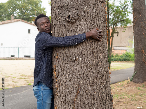 Primer plano de chico joven de raza negra, posando para una sesión fotográfica de retrato al aire libre, con camisa , abrazando un árbol .