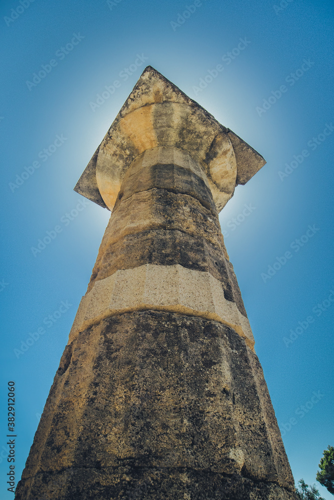 A vertical photo of Doric order column or pillar, photo taken on the ancient archeological site of Olympia, Greece, on a sunny day with sun shining directly behind the column