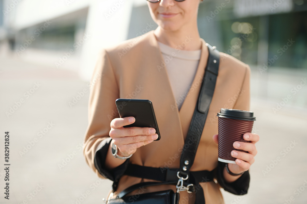 Hands of young businesswoman with glass of coffee scrolling through contacts