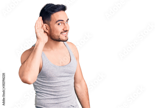 Young handsome man wearing swimwear and sleeveless t-shirt smiling with hand over ear listening an hearing to rumor or gossip. deafness concept.