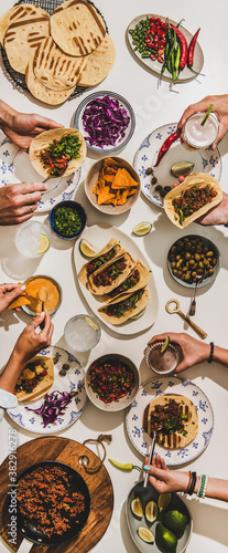 Friends having Mexican Taco dinner. Flat-lay of beef tacos, tomato salsa, tortillas, beer, snacks and peoples hands with food over white table, top view. Mexican cuisine, comfort food concept