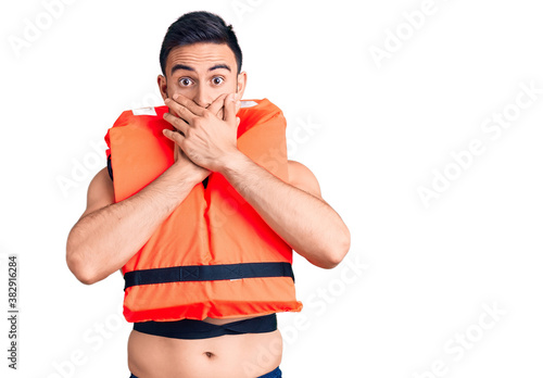 Young handsome man wearing lifejacket shocked covering mouth with hands for mistake. secret concept.