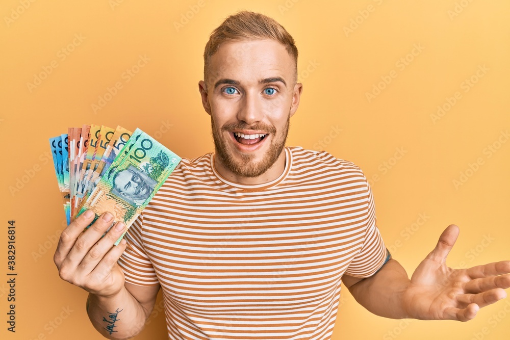 Young caucasian man holding australian dollars celebrating achievement with happy smile and winner expression with raised hand Young caucasian man holding australian dollars celebrating achievement with happy smile and winner expression with raised hand