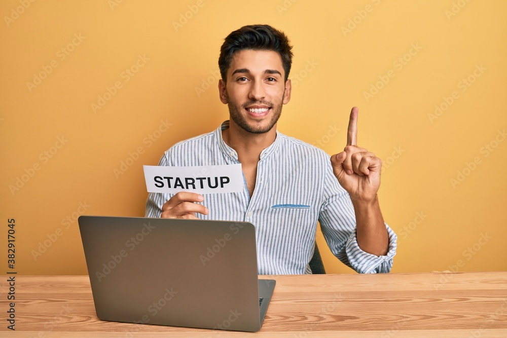 Young handsome man holding startup message as marketing development smiling with an idea or question pointing finger with happy face, number one Young handsome man holding startup message as marketing development smiling with an idea or question pointing finger with happy face, number one