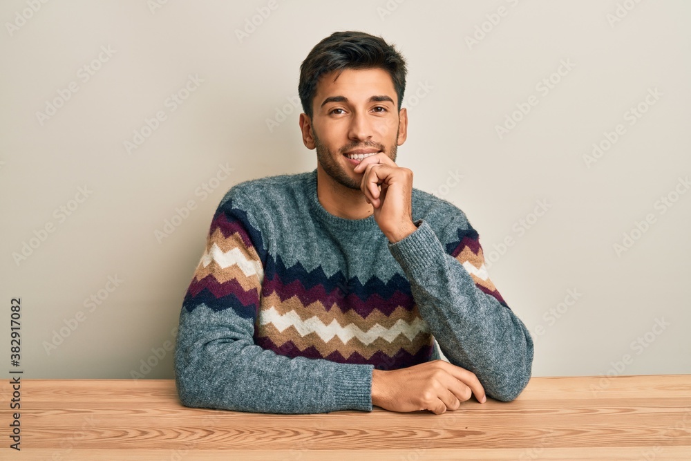 Young handsome man wearing casual sweater sitting on the table looking confident at the camera with smile with crossed arms and hand raised on chin. thinking positive. Young handsome man wearing casual sweater sitting on the table looking confident at the camera with smile with crossed arms and hand raised on chin. thinking positive.