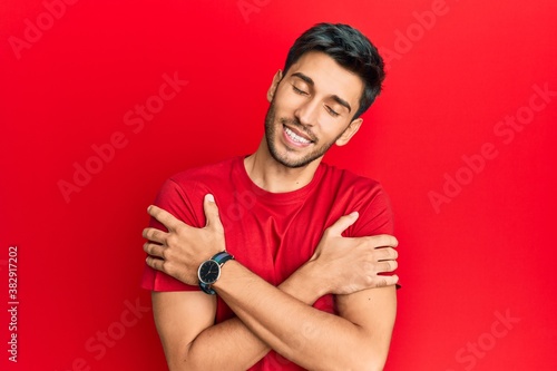 Fotografija Young handsome man wearing casual tshirt over red background hugging oneself happy and positive, smiling confident