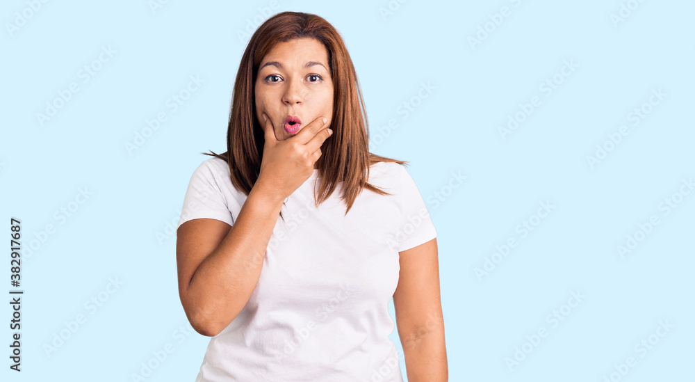 Middle age latin woman wearing casual white tshirt looking fascinated with disbelief, surprise and amazed expression with hands on chin Middle age latin woman wearing casual white tshirt looking fascinated with disbelief, surprise and amazed expression with hands on chin