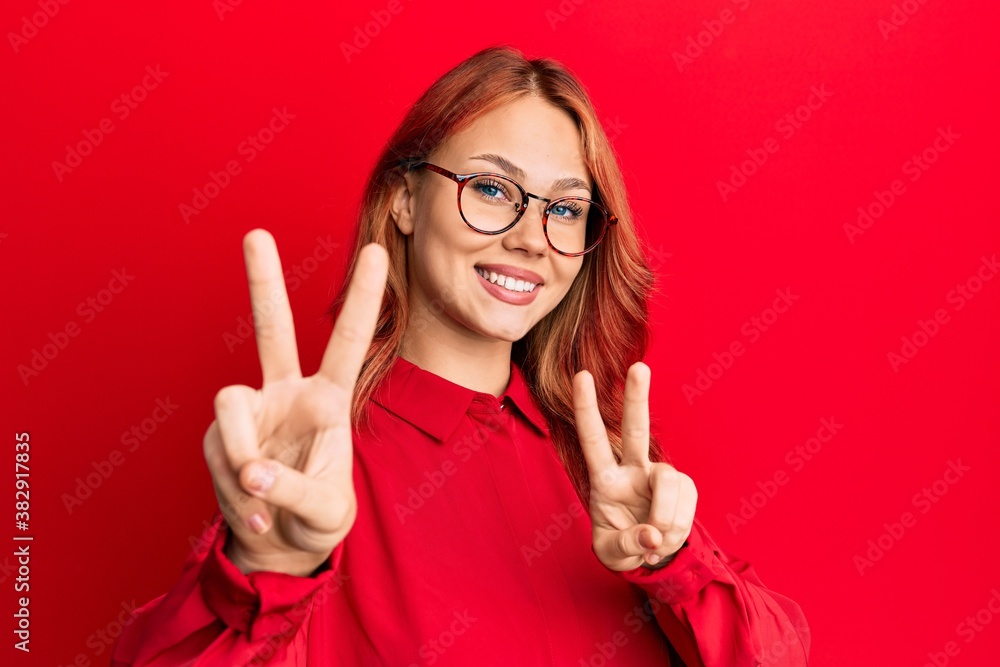 Young beautiful redhead woman wearing casual clothes and glasses over red background smiling looking to the camera showing fingers doing victory sign. number two. Young beautiful redhead woman wearing casual clothes and glasses over red background smiling looking to the camera showing fingers doing victory sign. number two.