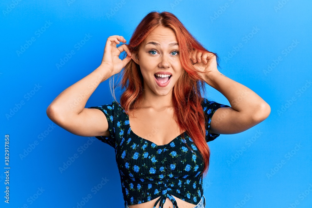 Young beautiful redhead woman wearing casual clothes over blue background smiling pulling ears with fingers, funny gesture. audition problem Young beautiful redhead woman wearing casual clothes over blue background smiling pulling ears with fingers, funny gesture. audition problem