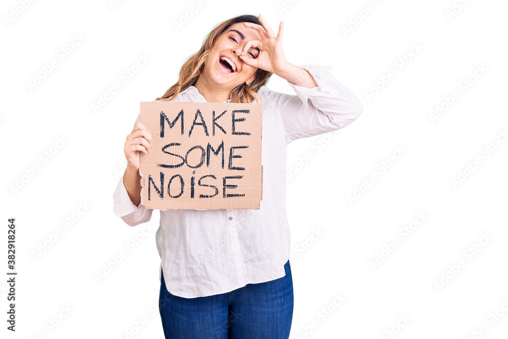 Young caucasian woman holding make some noise banner smiling happy doing ok sign with hand on eye looking through fingers Young caucasian woman holding make some noise banner smiling happy doing ok sign with hand on eye looking through fingers