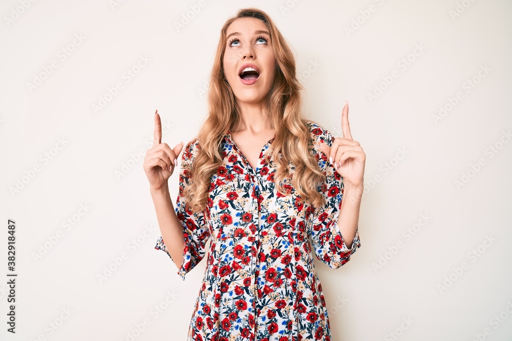Young caucasian woman with blond hair wearing summer dress amazed and surprised looking up and pointing with fingers and raised arms. Young caucasian woman with blond hair wearing summer dress amazed and surprised looking up and pointing with fingers and raised arms.