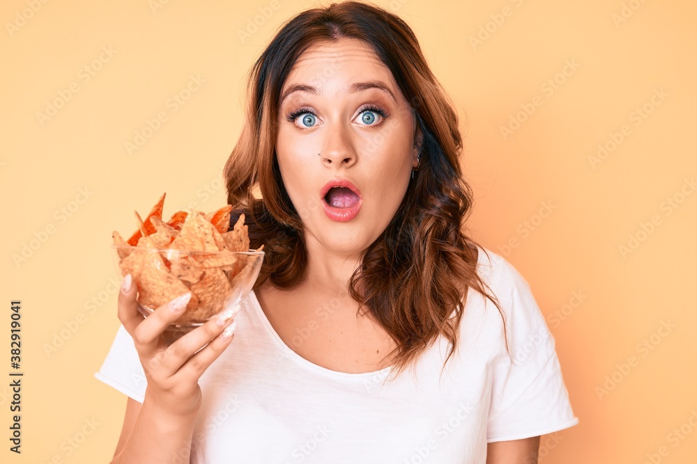 Young beautiful caucasian woman holding nachos potato chips scared and amazed with open mouth for surprise, disbelief face
