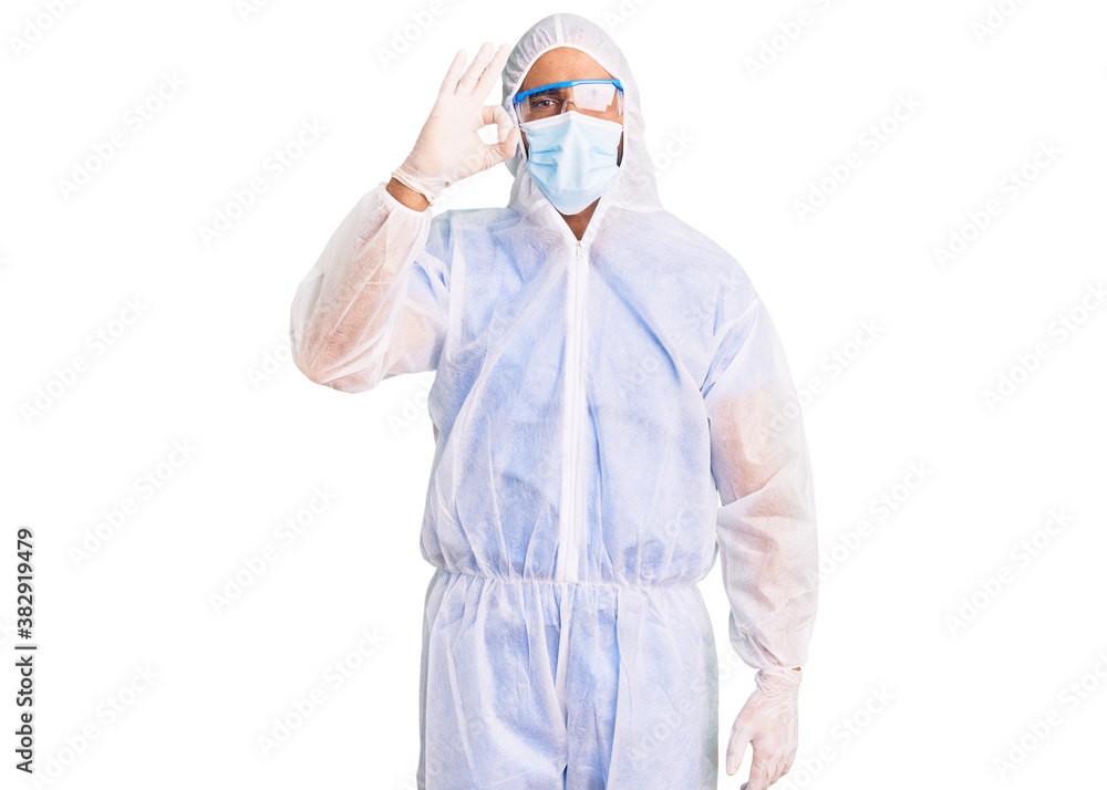 Young hispanic man wearing doctor protection coronavirus uniform and medical mask smiling positive doing ok sign with hand and fingers. successful expression. Young hispanic man wearing doctor protection coronavirus uniform and medical mask smiling positive doing ok sign with hand and fingers. successful expression.