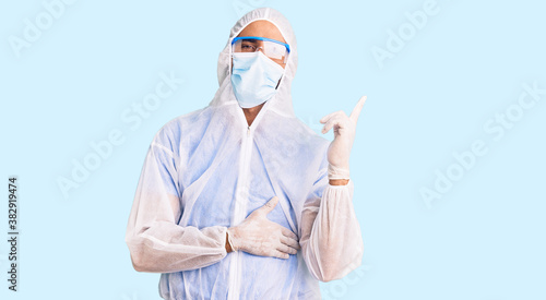 Young hispanic man wearing doctor protection coronavirus uniform and medical mask with a big smile on face, pointing with hand and finger to the side looking at the camera.