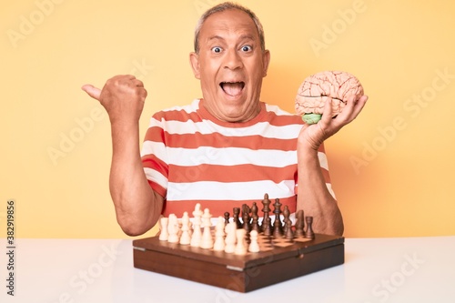 Senior handsome man with gray hair holding brain while playing chess pointing thumb up to the side smiling happy with open mouth