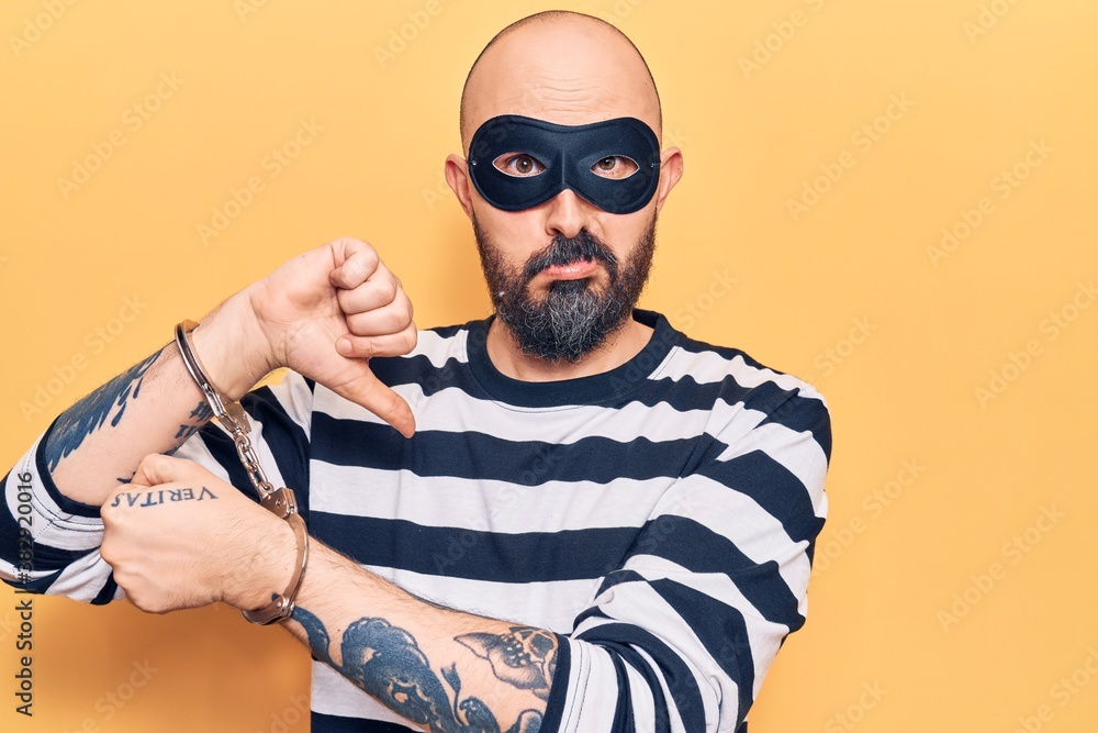 Young handsome man wearing burglar mask and handcuffs with angry face, negative sign showing dislike with thumbs down, rejection concept Young handsome man wearing burglar mask and handcuffs with angry face, negative sign showing dislike with thumbs down, rejection concept