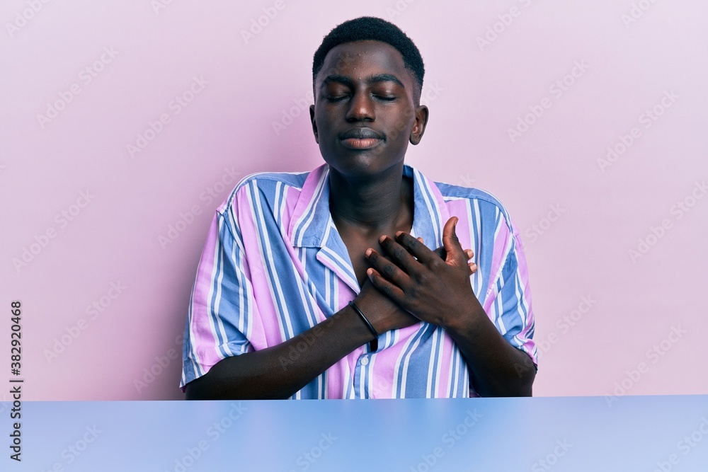 Young african american man wearing casual clothes sitting on the table smiling with hands on chest with closed eyes and grateful gesture on face. health concept. Young african american man wearing casual clothes sitting on the table smiling with hands on chest with closed eyes and grateful gesture on face. health concept.