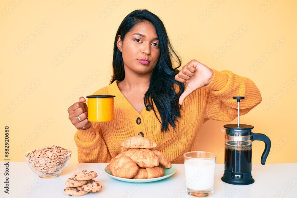 Beautiful latin young woman with long hair sitting on the table having breakfast with angry face, negative sign showing dislike with thumbs down, rejection concept Beautiful latin young woman with long hair sitting on the table having breakfast with angry face, negative sign showing dislike with thumbs down, rejection concept