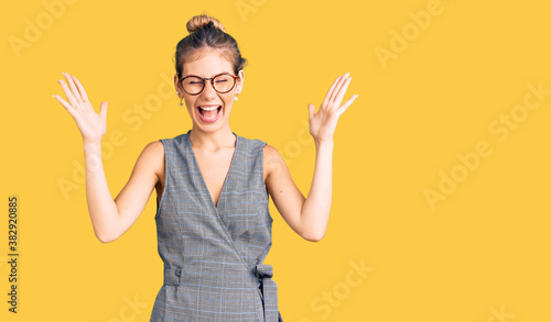 Beautiful caucasian woman with blonde hair wearing business clothes and glasses celebrating mad and crazy for success with arms raised and closed eyes screaming excited. winner concept