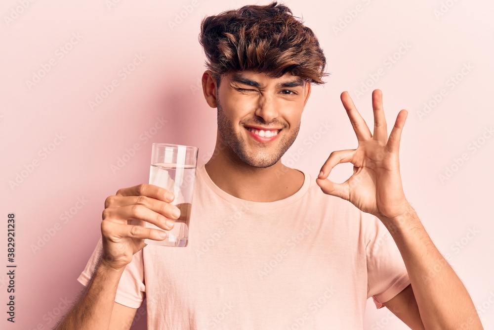 Young hispanic man drinking glass of water doing ok sign with fingers, smiling friendly gesturing excellent symbol Young hispanic man drinking glass of water doing ok sign with fingers, smiling friendly gesturing excellent symbol
