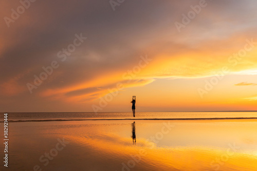 Silhouette of carefree fitness woman jumping on the beach at sunset, raising her hands up. Freedom concept. 