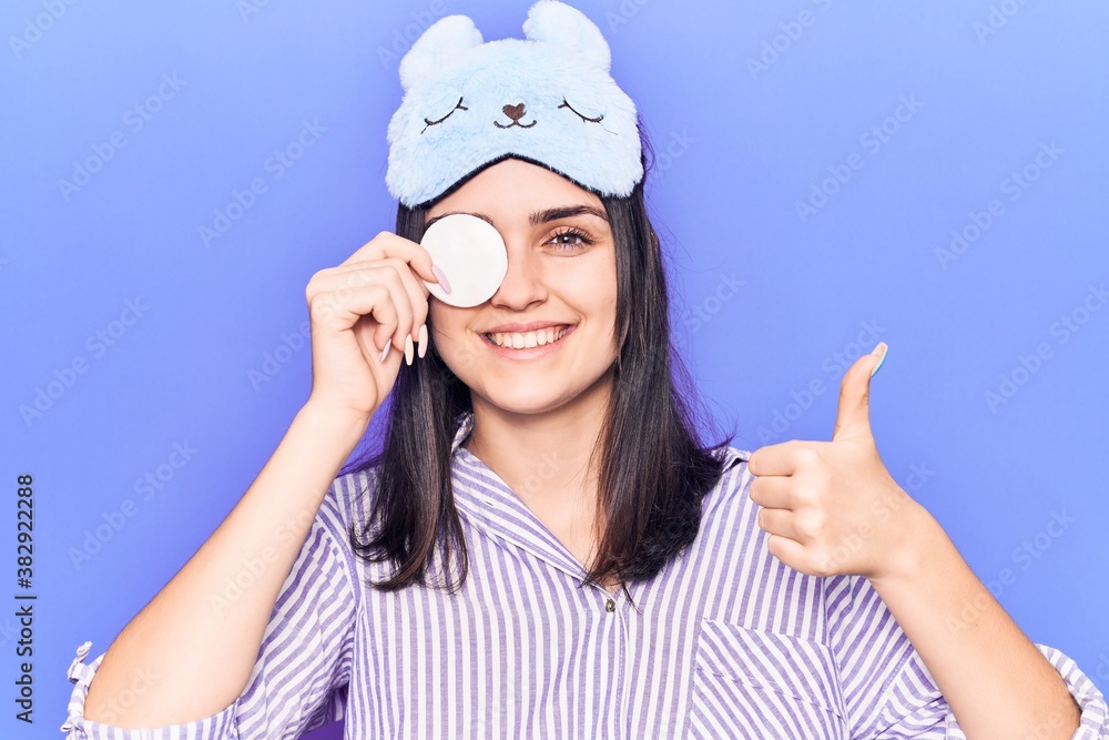 Young beautiful girl wearing funny sleep mask using makeup remover cotton smiling happy and positive, thumb up doing excellent and approval sign Young beautiful girl wearing funny sleep mask using makeup remover cotton smiling happy and positive, thumb up doing excellent and approval sign