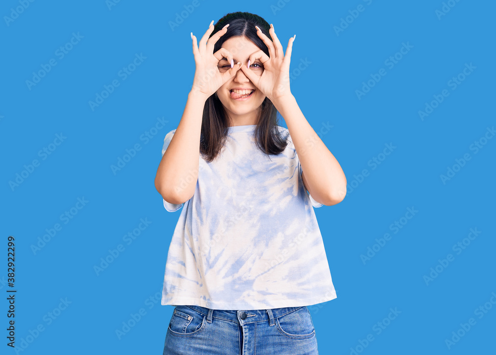 Young beautiful girl wearing casual t shirt doing ok gesture like binoculars sticking tongue out, eyes looking through fingers. crazy expression. Young beautiful girl wearing casual t shirt doing ok gesture like binoculars sticking tongue out, eyes looking through fingers. crazy expression.